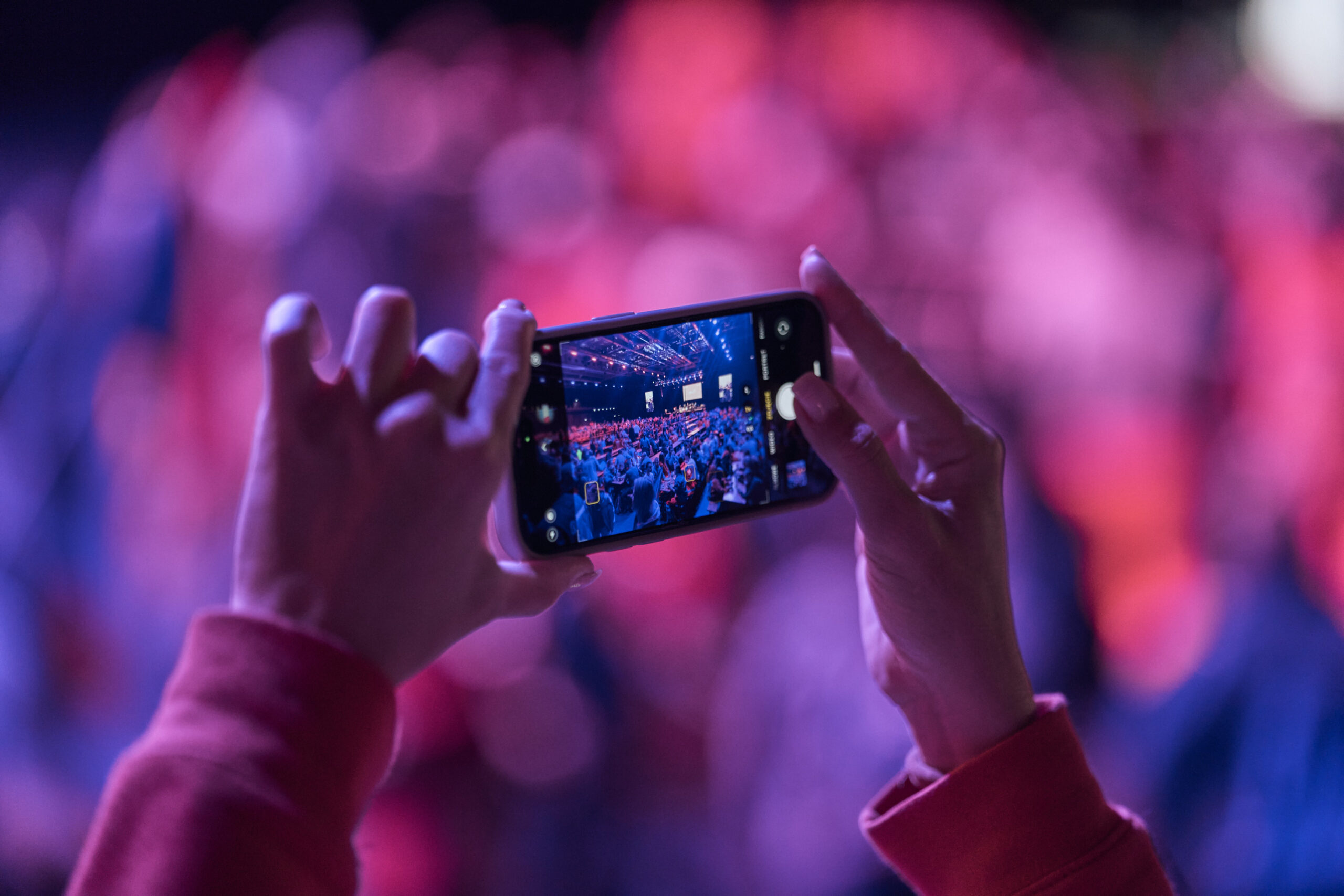 Jemand nimmt ein Erinnerungsfoto von der Veranstaltung mit einem Smartphone auf. Zwei Hände halten das Telefon, der Hintergrund ist in blauen und roten Farbtönen.