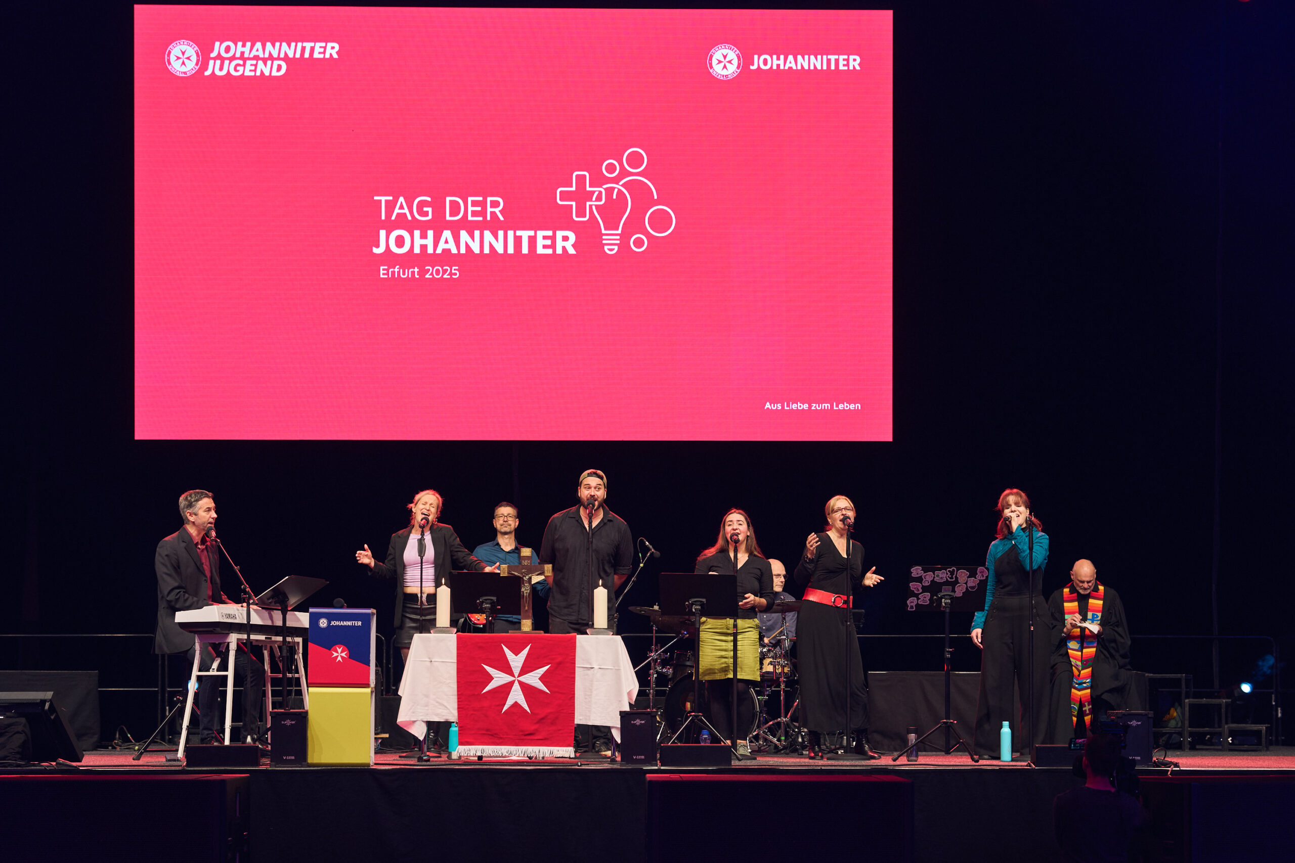 Auf einer Bühne stehen Menschen, die gemeinsam singen und Musik machen. Im Hintergrund sieht man einen großen Bildschirm mit der Schrift: Tag der Johanniter Erfurt 2025.