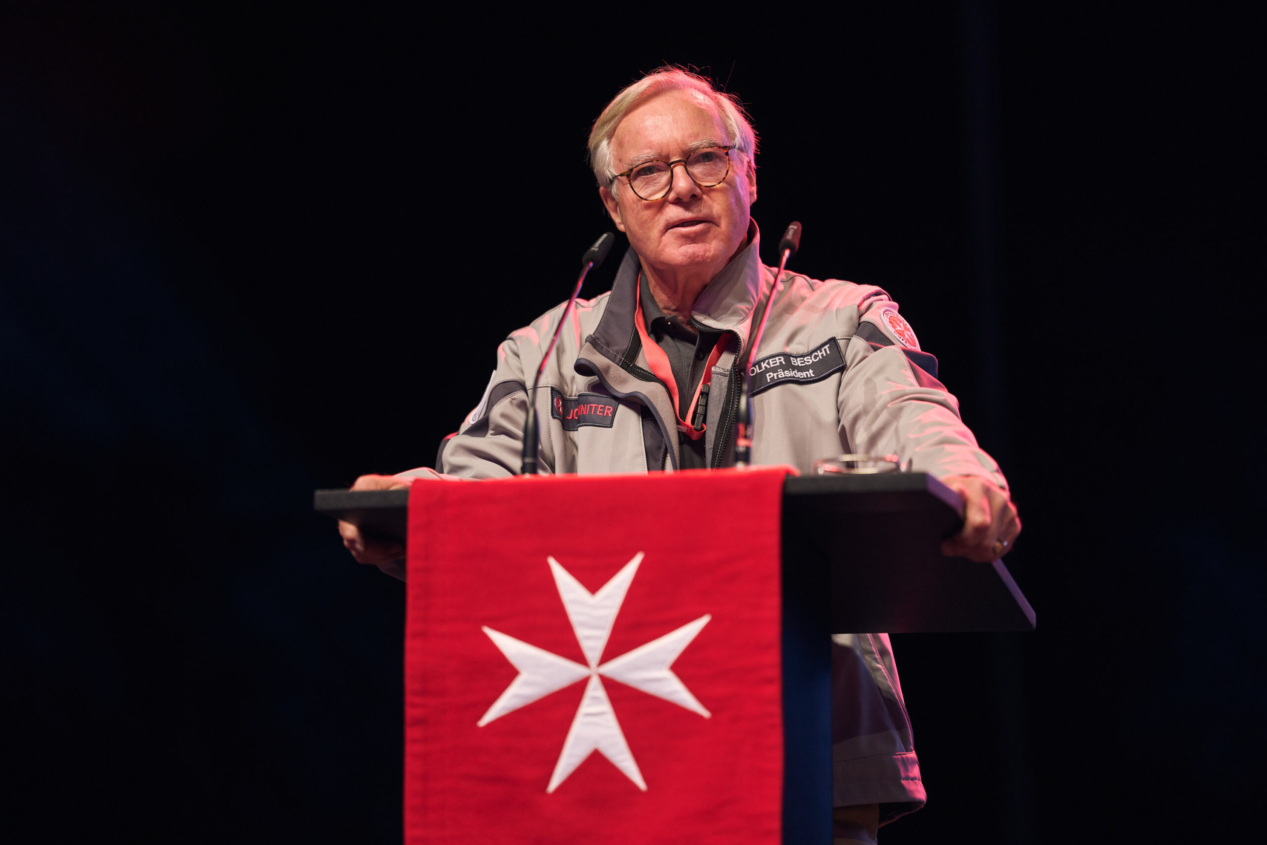 Man sieht einen Mann, der an einem Redepult mit dem Zeichen der Johanniter steht und spricht. Das Logo ist ein weißes achtspitziges Kreuz auf roten Grund.