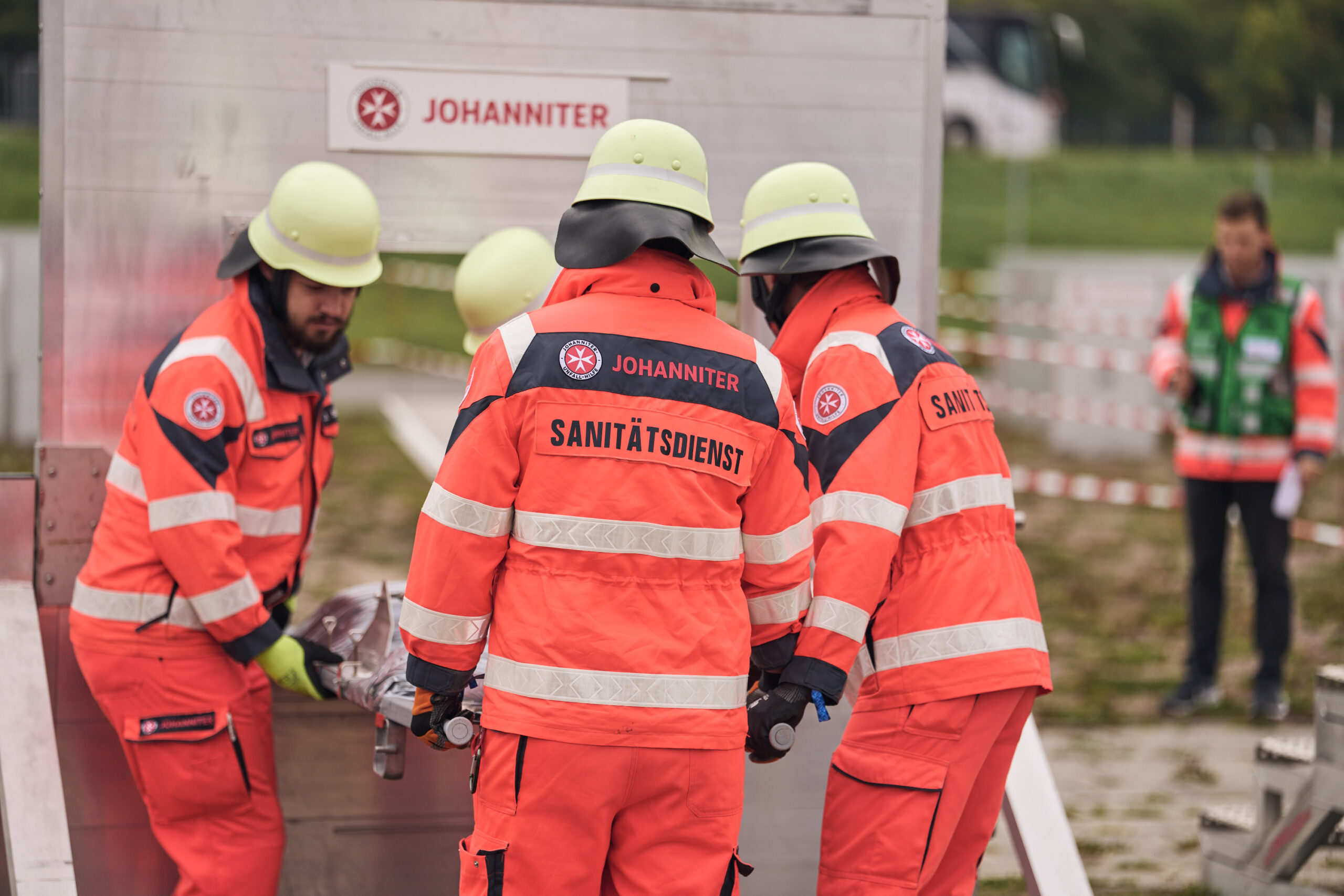 Vier Einsatzkräfte in Einsatzkleidung heben eine Trage durch einen Hindernisparcours. Auf der orangenen Einsatzkleidung steht das Wort: Sanitätsdienst. Im Hintergrund ist ein Schiedsrichter zu erkennen.