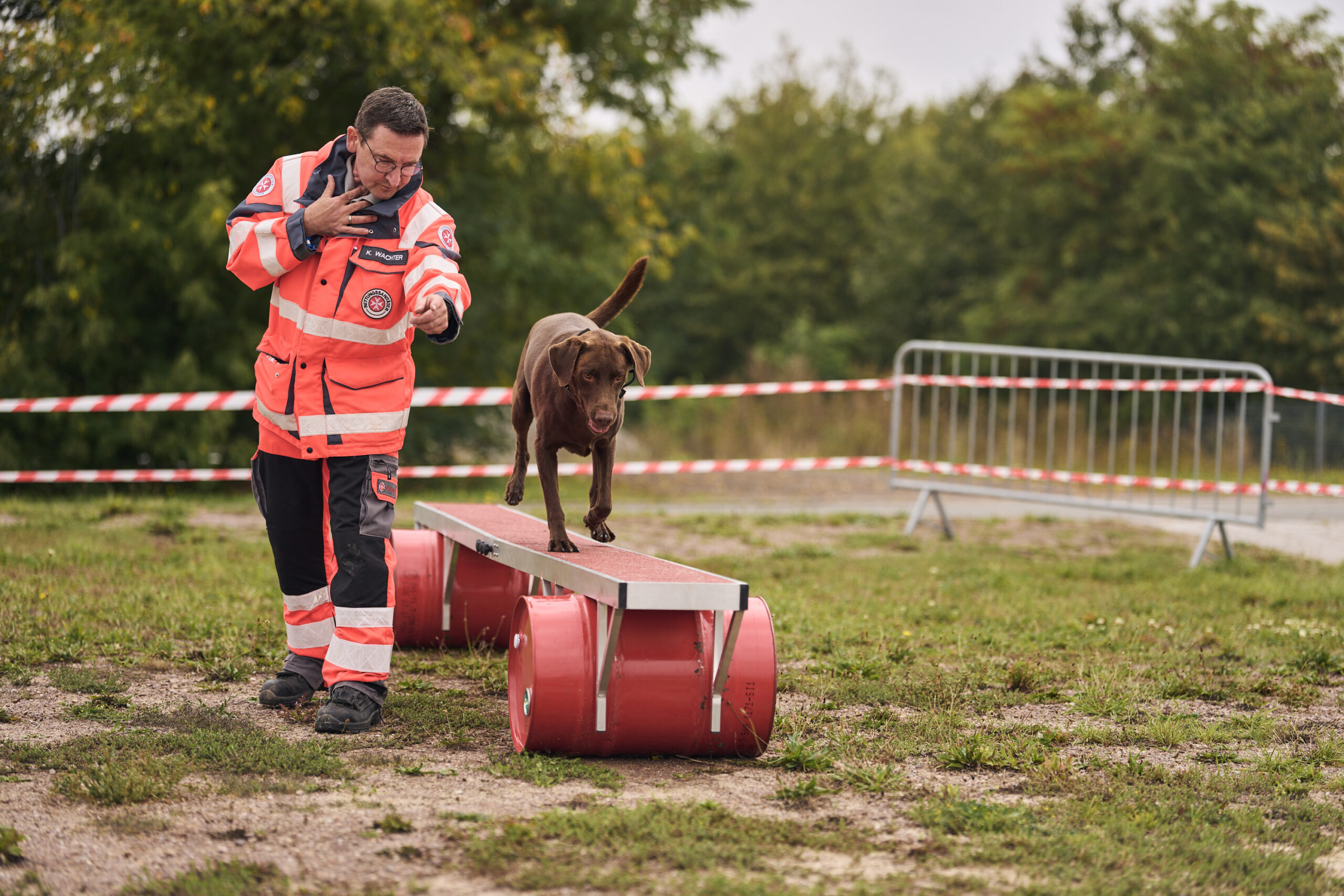 Ein Mann in der Einsatzkleidung der Johanniter schickt einen braunen Hund über einen Parcours. Der Bereich ist durch ein rot-weißes Band und ein Gitter eingegrenzt und im Hintergrund sieht man grüne Natur.