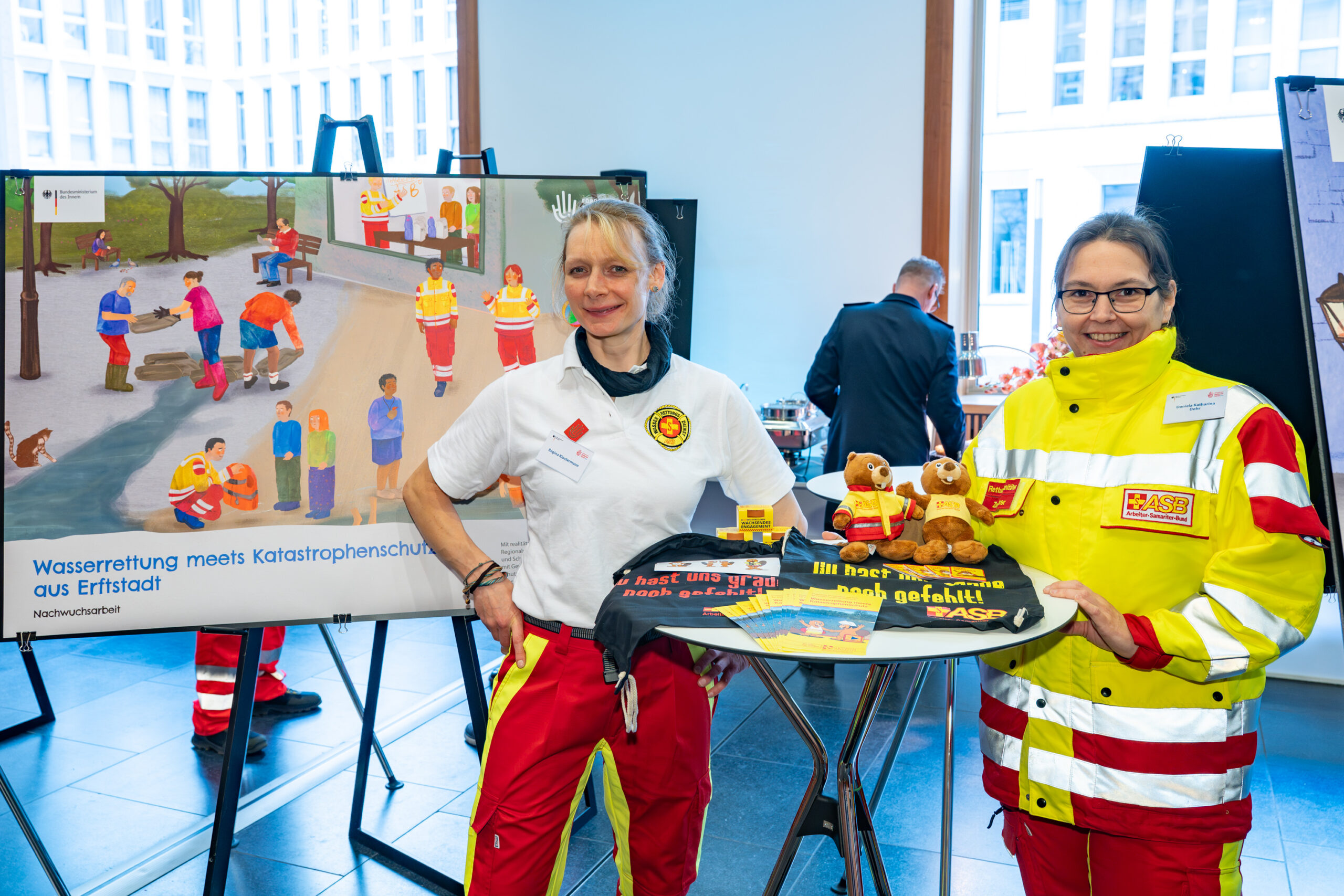 Zwei Einsatzkräfte in rot-gelber Uniform stellen bei einer Ausstellung ihr Projekt vor.